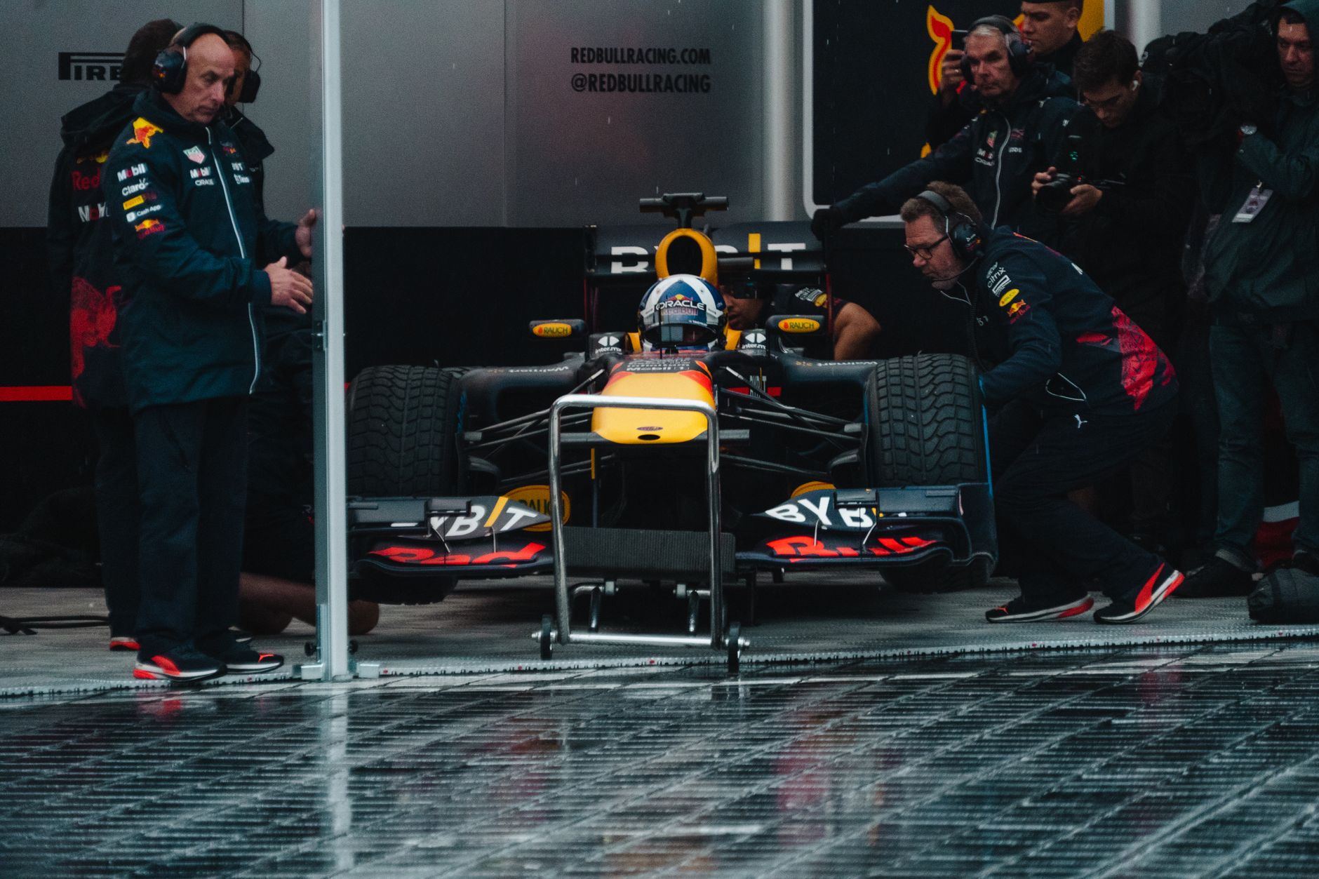 Monoplace de Formule 1 dans un garage sous la pluie, pilote casqué à bord, entourée de mécaniciens en tenue d’équipe et de spectateurs.