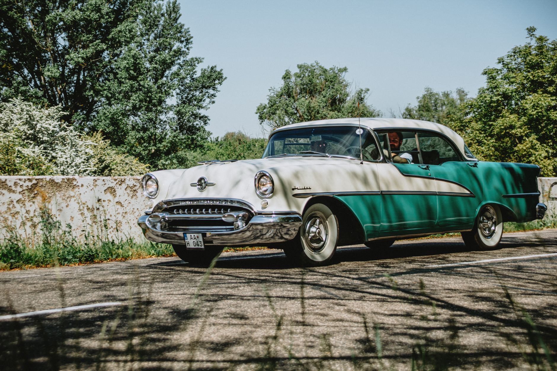 Voiture ancienne blanche et verte roulant sur une route bordée d’un muret et d’arbres, par temps clair.